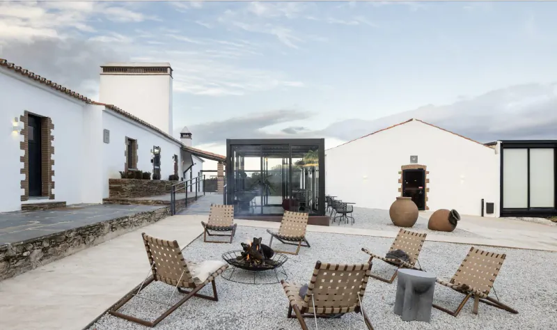 Modern outdoor patio in Alandroal with white buildings, glass pavilion, rattan chairs around fire pit, large pots, cloudy sky.