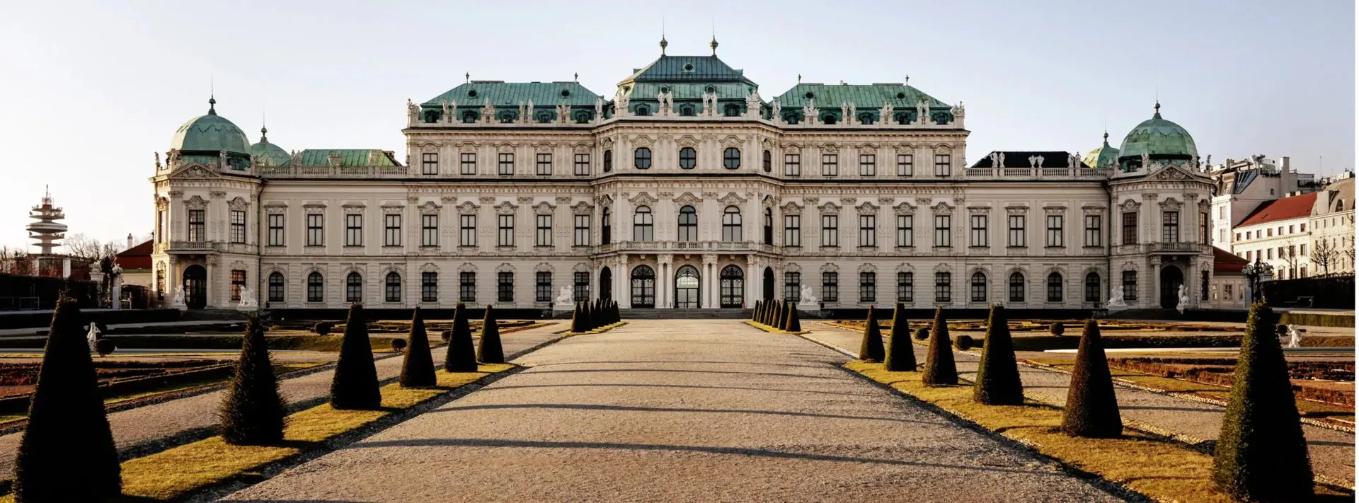 Belvedere Palace in Vienna, grand white Baroque facade with green domes, flanked by manicured topiary trees on a pathway.