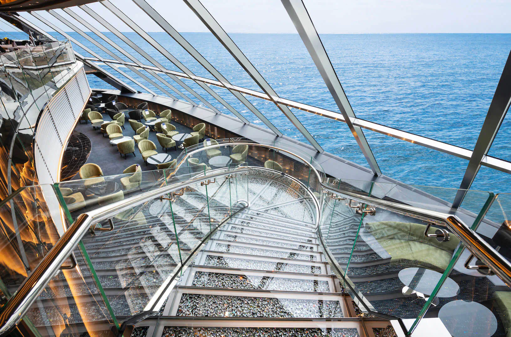 Glass spiral staircase in MSC Virtuosa Yacht Club lounge overlooking ocean, with curved seating and tables.