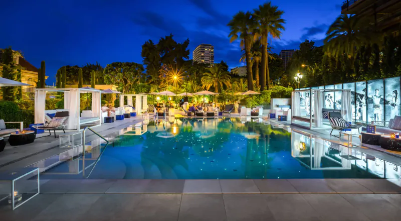 Twilight pool at Hotel Metropole Monte-Carlo: glowing blue water, white cabanas, palm trees, city lights.