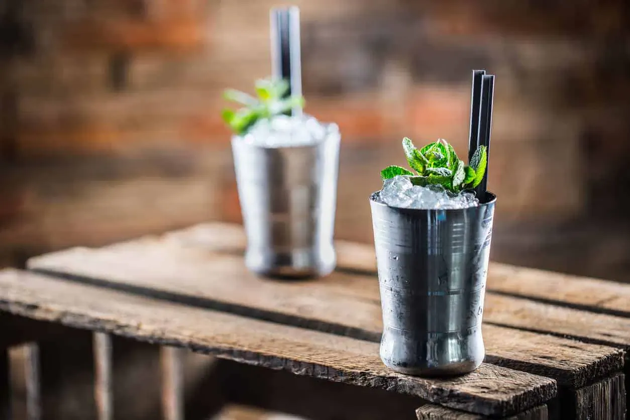 Two mint juleps in silver cups garnished with mint sprigs and straws on wooden table against brick wall.