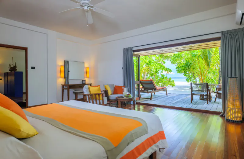 Spacious beachfront hotel room with orange bed, chairs, open doors to tropical ocean view at Mirihi Island Resort