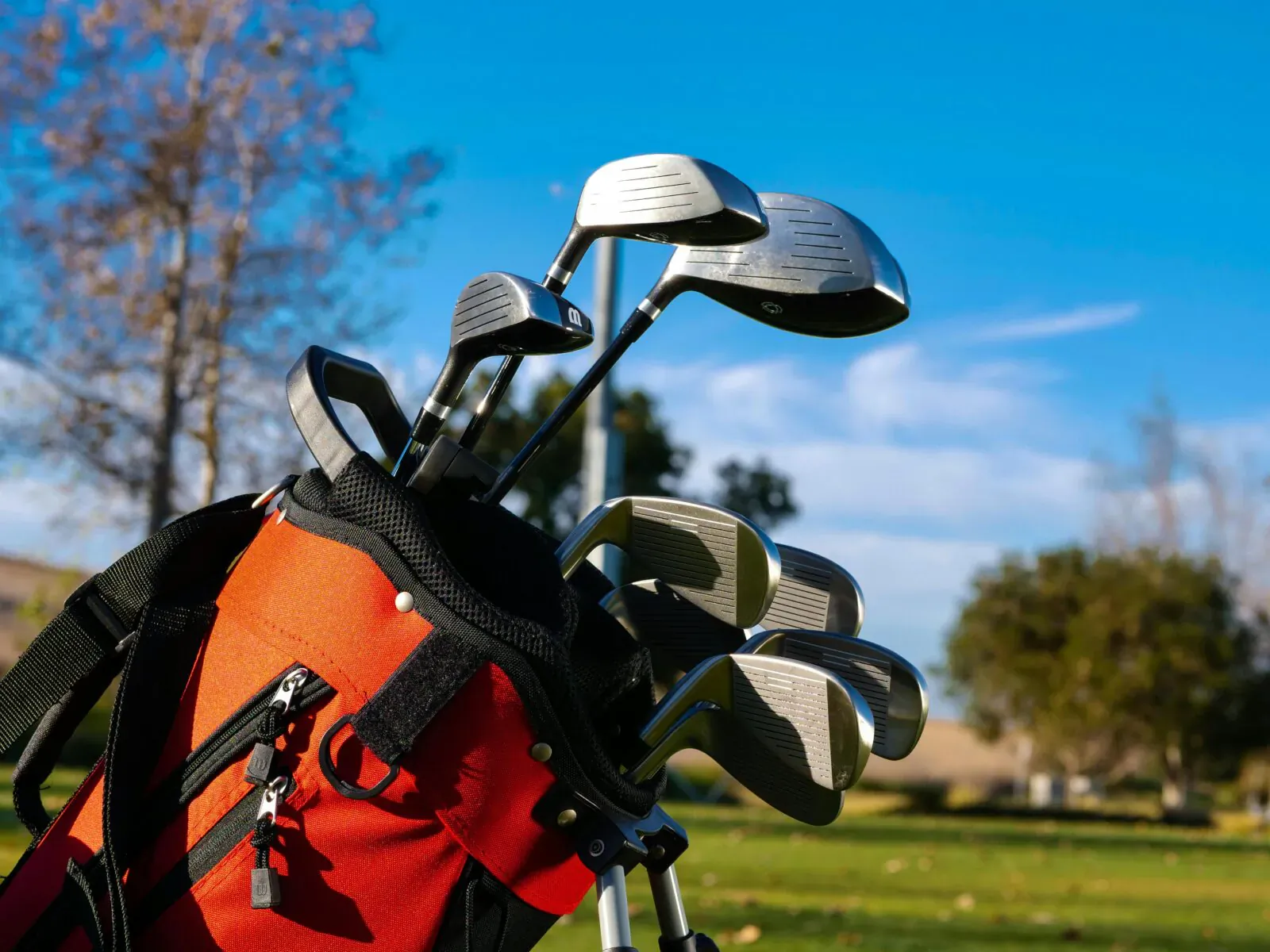 Orange golf bag with silver hybrid clubs outdoors on green field under blue sky