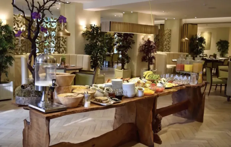 Luxurious hotel breakfast buffet on wooden table with fruits, pastries, juices amid elegant indoor plants and seating.