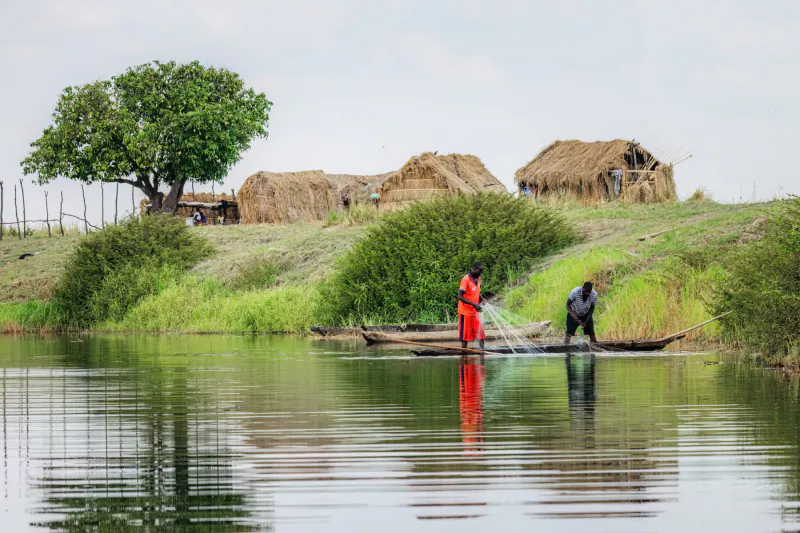 Woman in red and man in black fishing from dugout canoe on calm lake, thatched huts and acacia tree nearby, Zambia.
