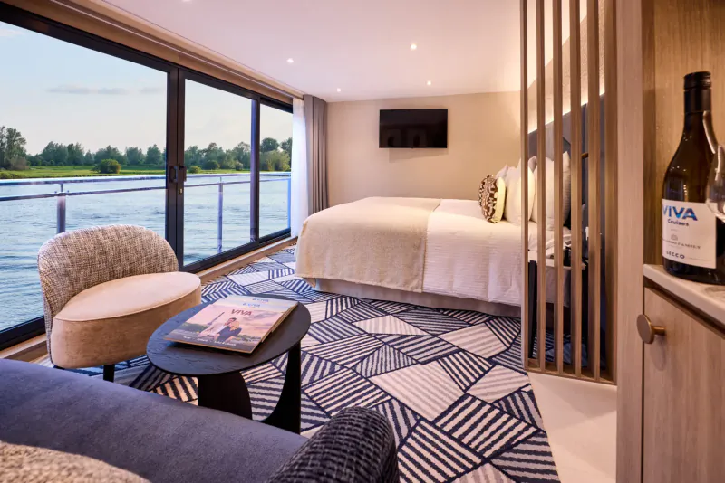 Luxurious river cruise suite with king bed, chairs, VIVA wine bottle, river view through panoramic windows.