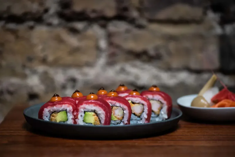 Row of red tuna sushi rolls topped with orange beads on black plate, avocado filling visible, ginger and chopsticks beside, rustic stone wall backdrop