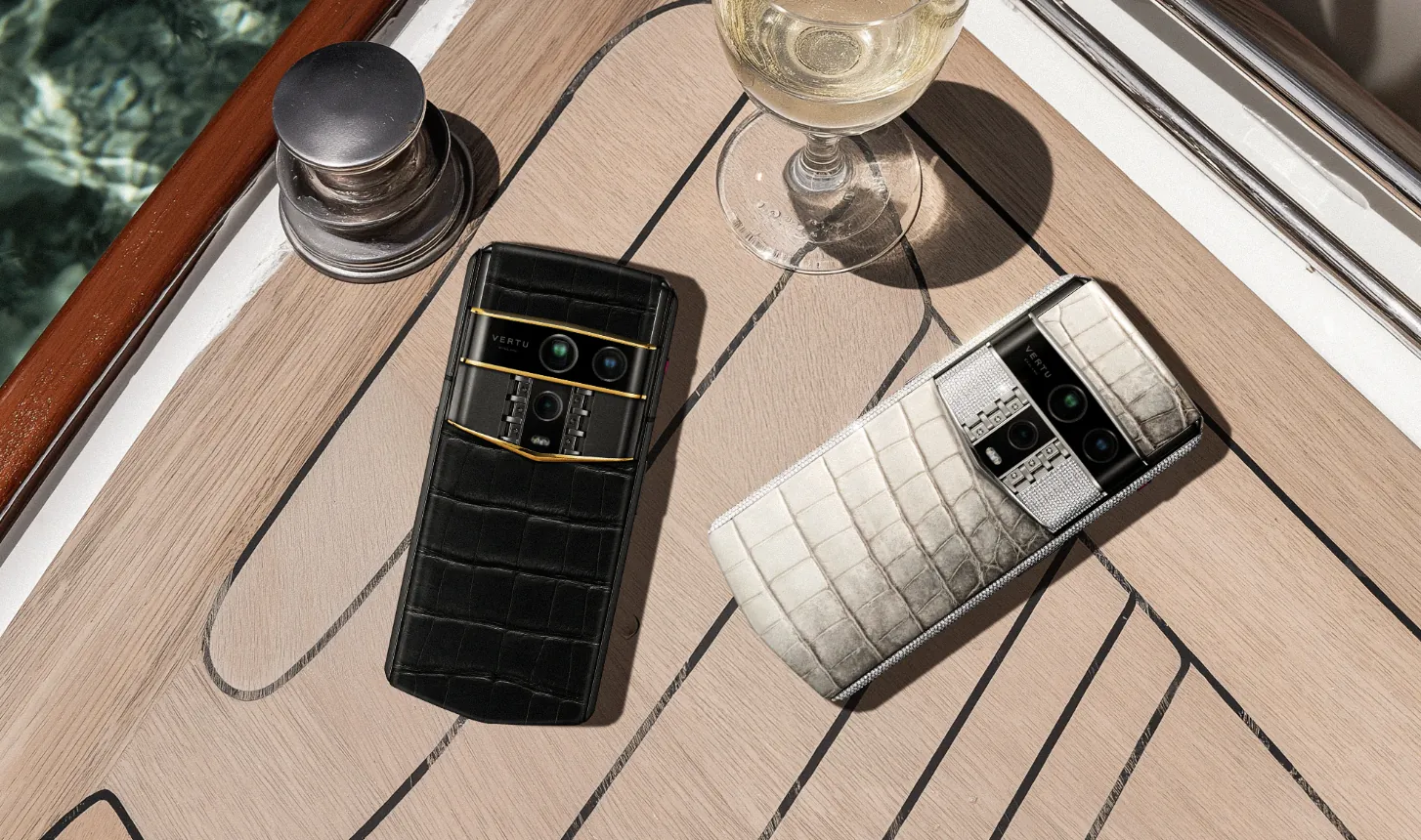 Black and white crocodile-textured Vertu phones beside wine glass on luxury yacht deck.