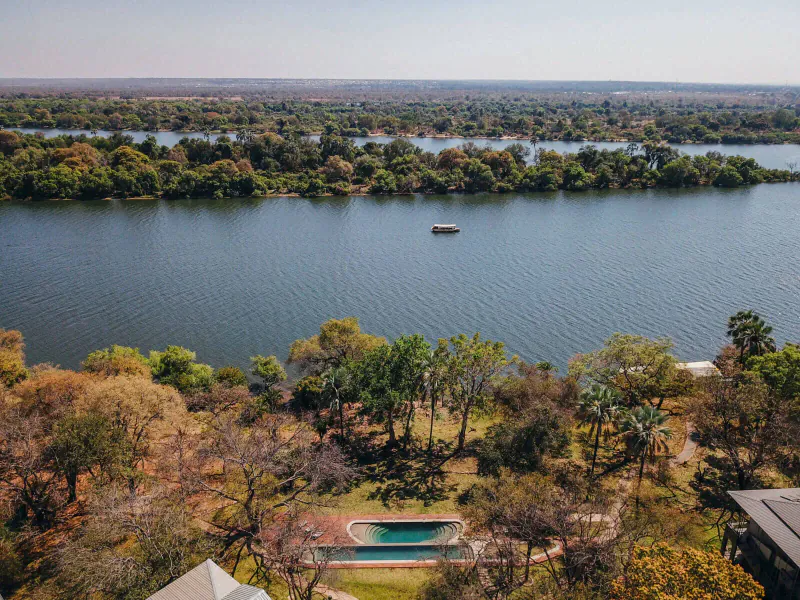 Aerial view of Palm River Hotel pool overlooking Zambezi River with distant Victoria Falls horizon and small boat.