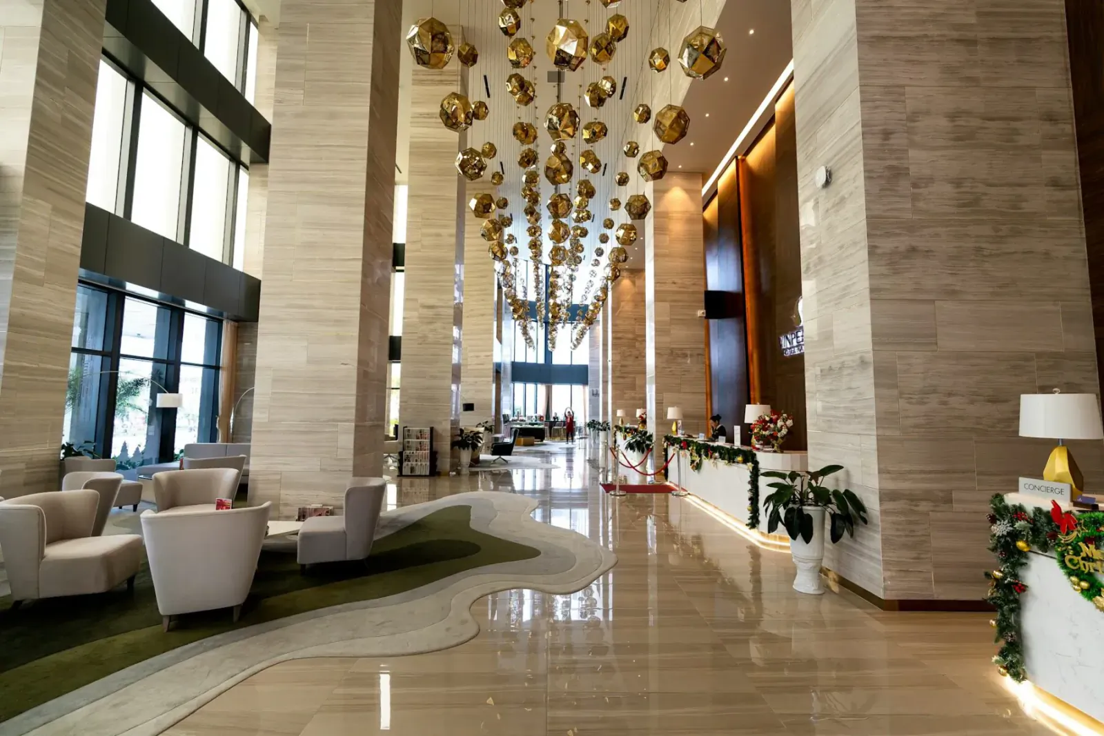Luxurious hotel lobby with golden orb chandeliers, beige stone columns, seating area, reception, holiday garlands, and polished floors.