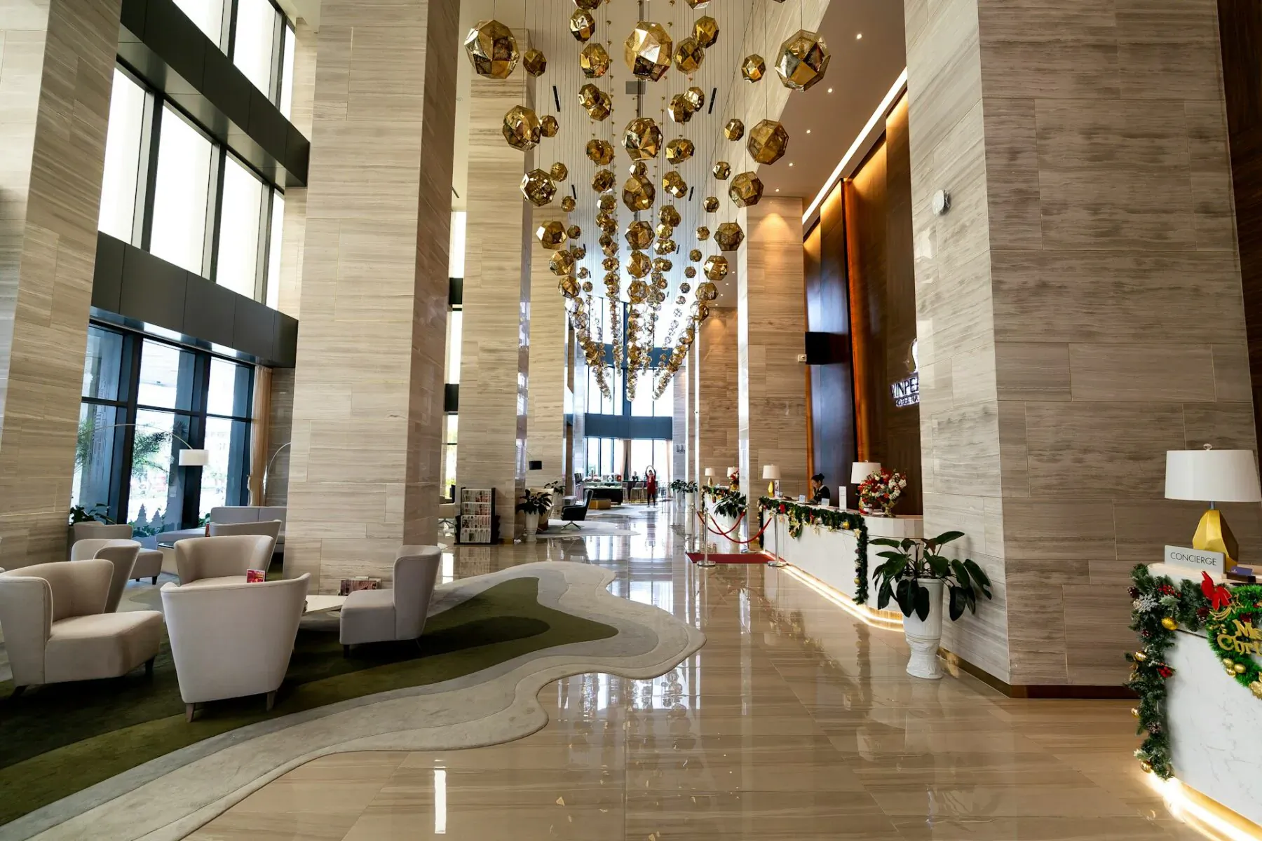 Luxurious hotel lobby with golden orb chandeliers, beige stone columns, seating area, reception, holiday garlands, and polished floors.