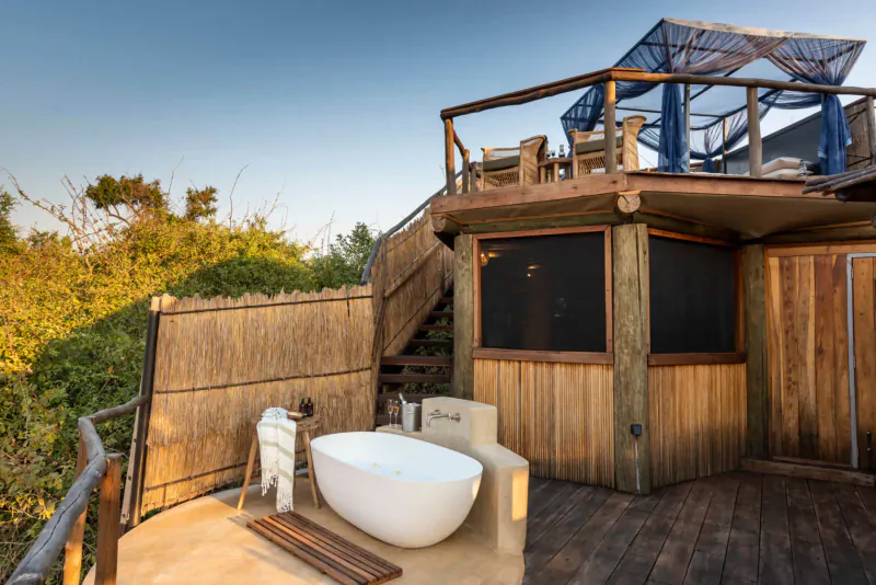Outdoor luxury bathtub on wooden deck of elevated thatched Lion Camp lodge amid savanna greenery at sunset