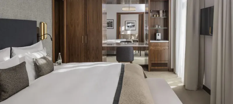 Luxurious hotel bedroom with king bed, wood wardrobe, desk, TV, and window in St. Moritz suite