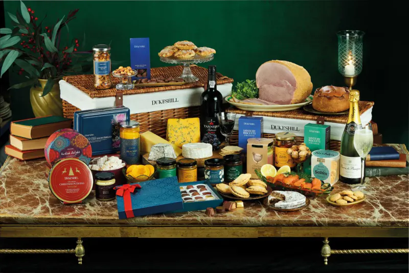 Festive Christmas hamper on ornate table: Dukeshill meats, cheeses, chocolates, wine, champagne amid greenery.