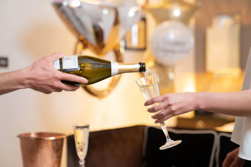 Man pouring champagne from bottle into woman's flute glass amid balloons and 'Congratulations' decor at Sheraton event