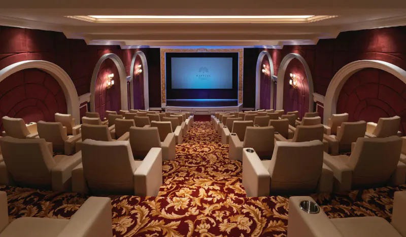 Luxurious empty cinema at Raffles The Palm Dubai with plush beige seats, red walls, ornate carpet, and lit screen.