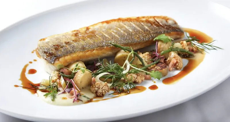 Pan-seared sea bass fillet on white oval plate with baby potatoes, chickpeas, herbs, and soy sauce drizzle.
