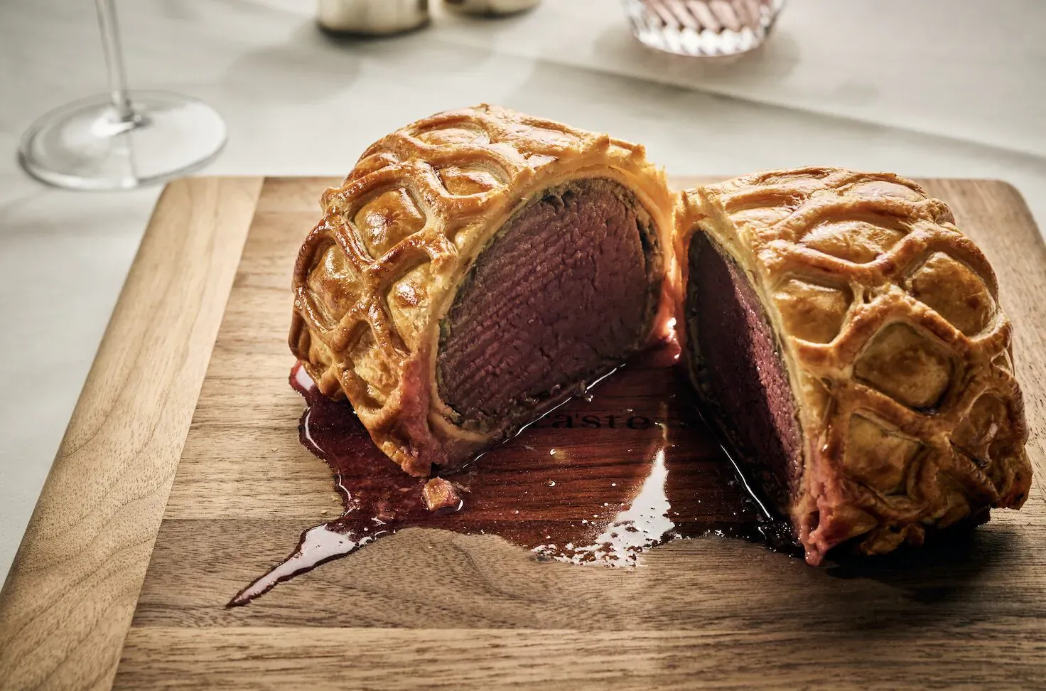 Beef Wellington sliced open on wooden board, revealing pink center and juices, with wine glass on white tablecloth