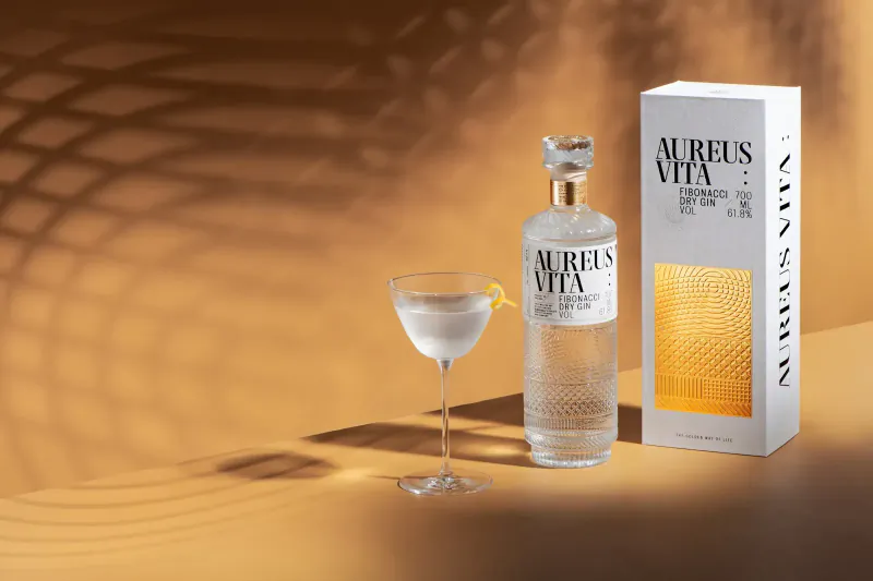 Aurel's Vita luxury gin bottle and glass of gin with lemon twist beside white box on warm backdrop.