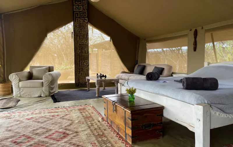 Spacious tent interior at Tumaren Camp with beige bed, trunk, armchair, and savanna view through windows.