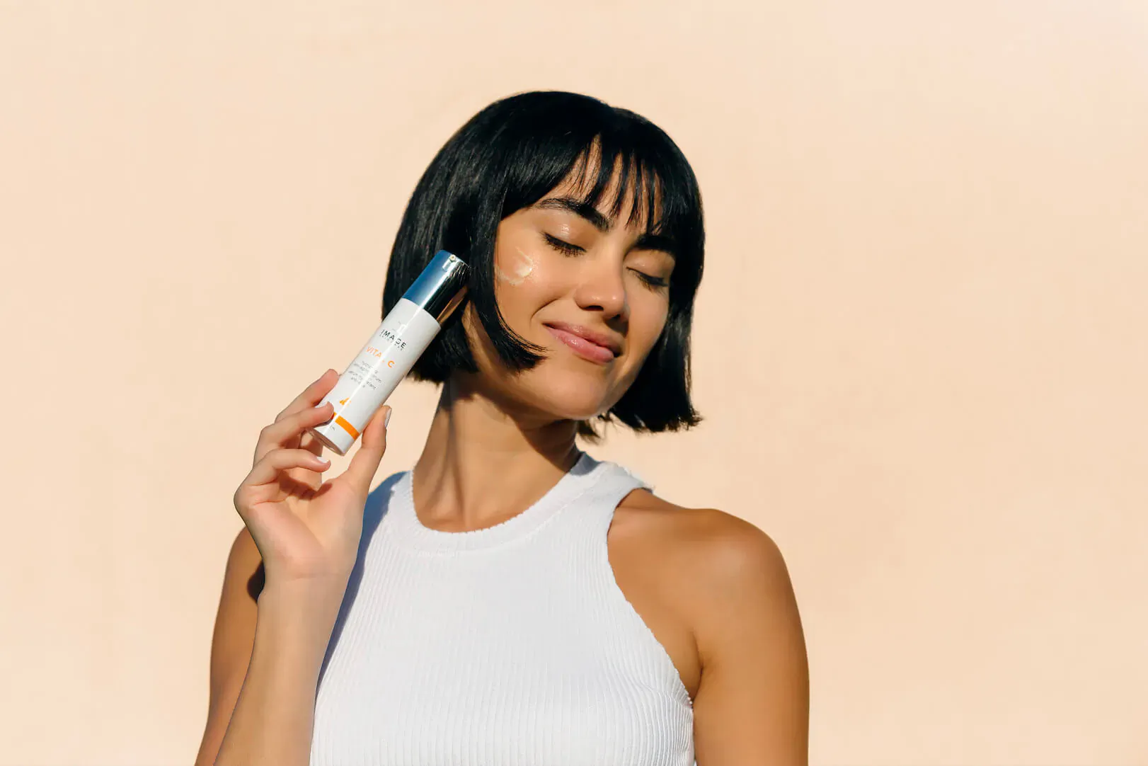 Smiling woman with bob haircut holds Image Skincare bottle to cheek, wearing white tank top on beige background