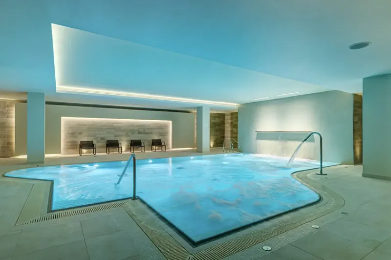 Modern indoor pool at Apex City of Bath Hotel with blue-lit water, lounge chairs, and tiled walls.