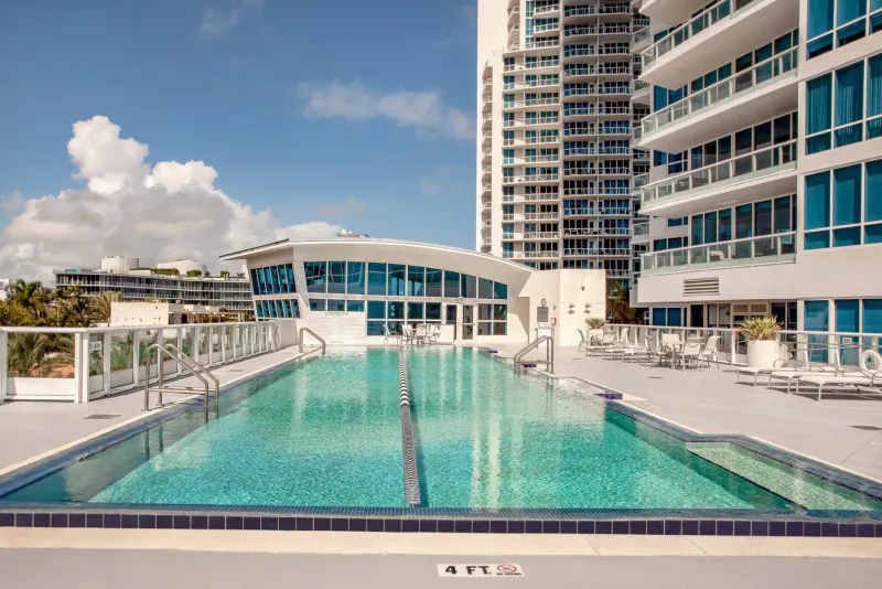 Infinity-edge pool at modern high-rise resort with curved glass buildings, lounge chairs, blue sky, and palm trees.