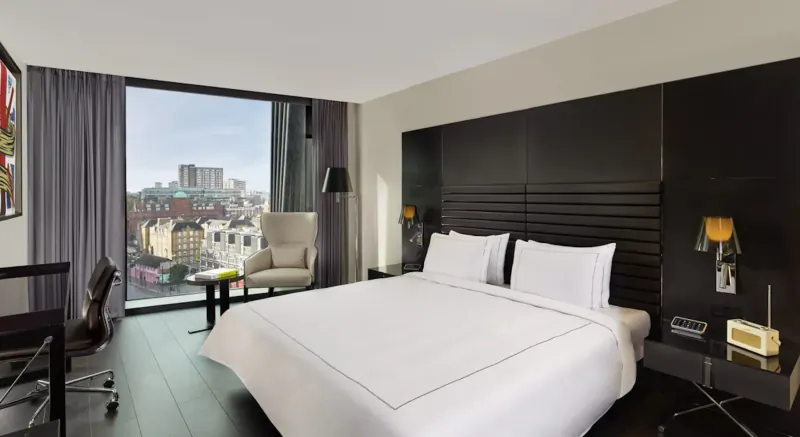 Modern hotel room at art'otel London Hoxton with king bed, armchair, desk, and city skyline view through large window.