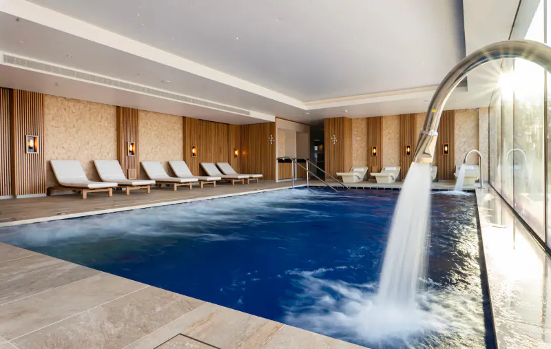 Luxury spa pool with cascading water from overhead shower, white loungers, wood panels, and large windows at Carden Park.