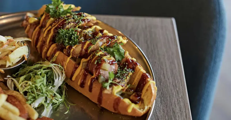 Close-up of a hot dog topped with sauce and parsley on a plate with coleslaw, on a wooden table.