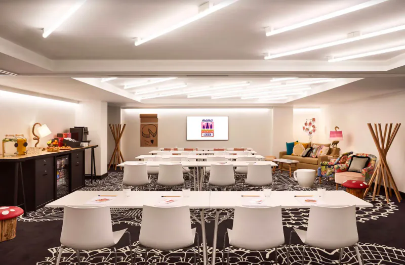 Modern Mama Shelter East London meeting room with white tables, chairs, bar, plants, and screen on wall.