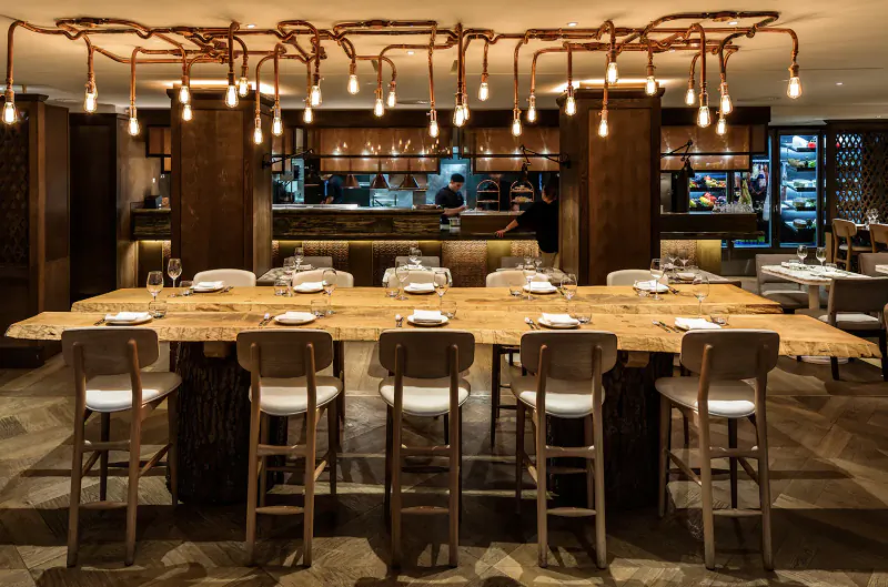 Elegant hotel dining area with live-edge wooden table, bar stools, pendant lights, and open kitchen.