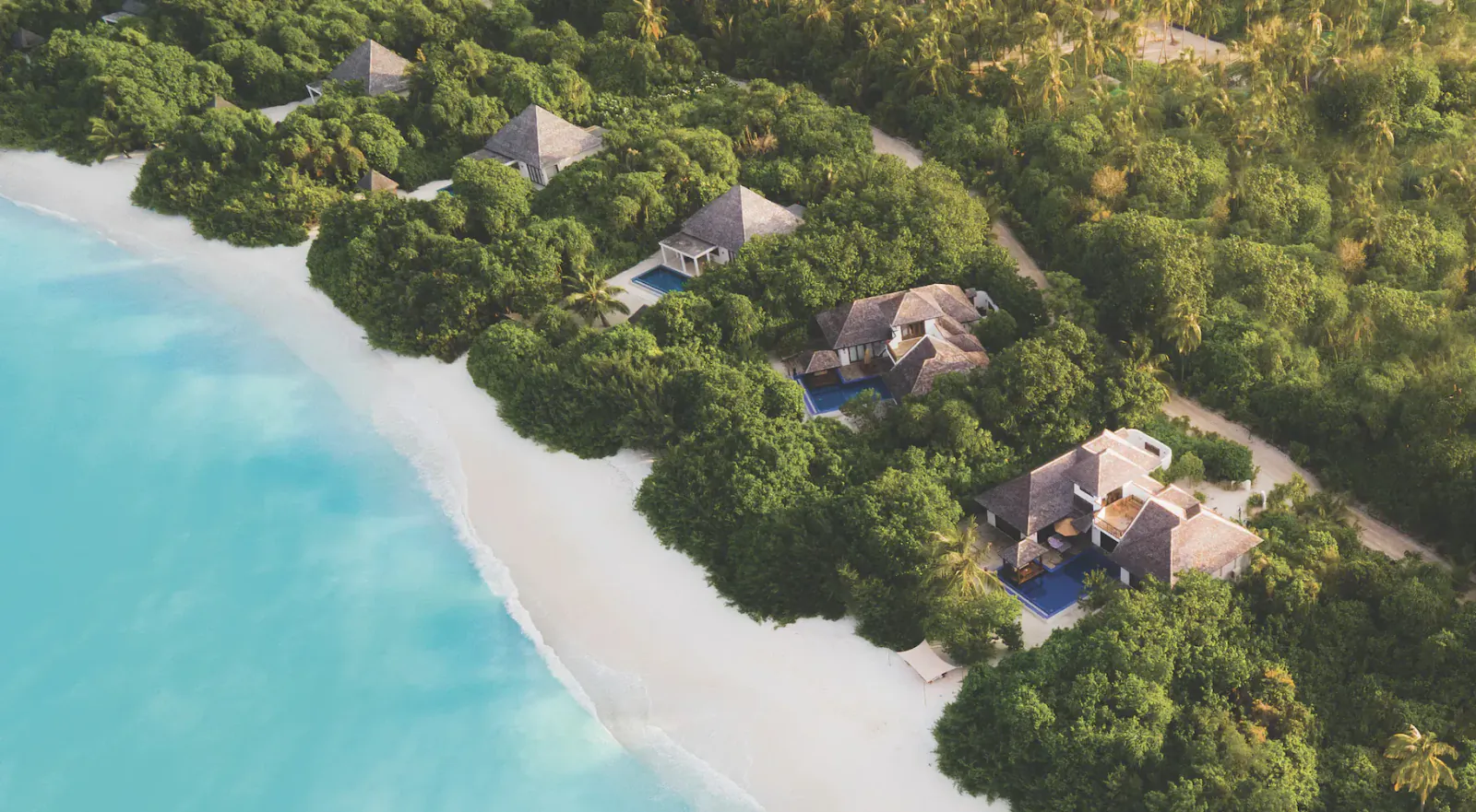 Aerial view of luxury thatched villas nestled in lush greenery along white-sand beach at Hideaway Beach Resort, Maldives, with turquoise ocean.