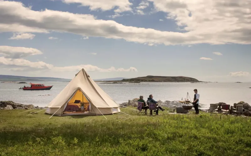 Beige bell tent on grassy shore by calm sea, people seated nearby with chairs and cooler, red boat and islands in background under cloudy sky