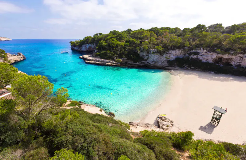 Aerial view of turquoise Cala Macarelleta beach in Menorca, surrounded by cliffs, pine trees, and a lifeguard tower.