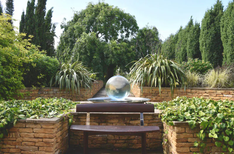 Large reflective glass sphere fountain on stone bench in lush garden with trees and greenery at Cotswold House Hotel & Spa