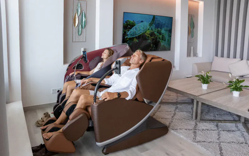 Man and woman relaxing in brown massage chairs in modern hotel lounge with ocean TV and plants.