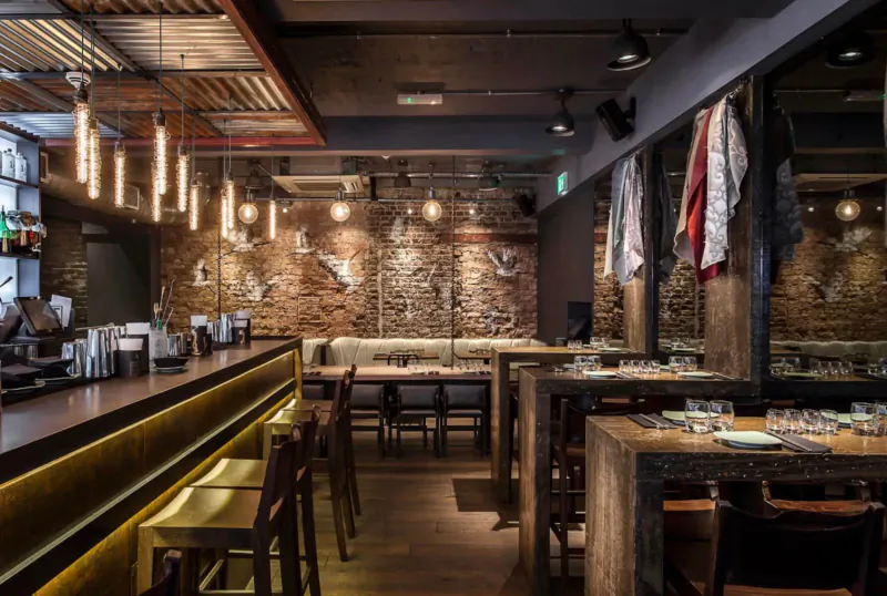 Modern industrial-style bar interior with brick walls, hanging lights, wooden tables, bar stools, and glassware.