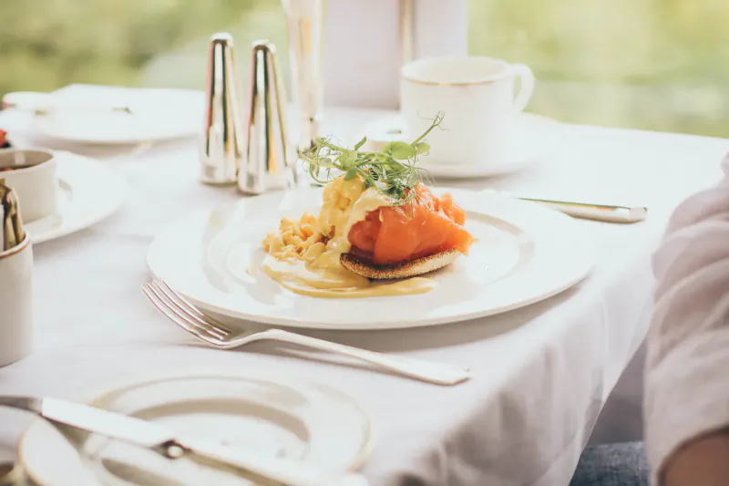 Luxury train dining table with poached salmon on eggs with hollandaise, garnished with herbs, white cup, salt and pepper shakers, window view.