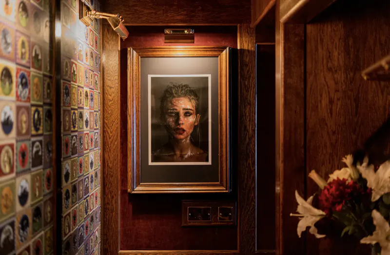 Framed portrait of a woman in ornate gold frame on a narrow, colorful wallpapered hallway wall at House of Gods Hotel gallery, with flowers nearby.