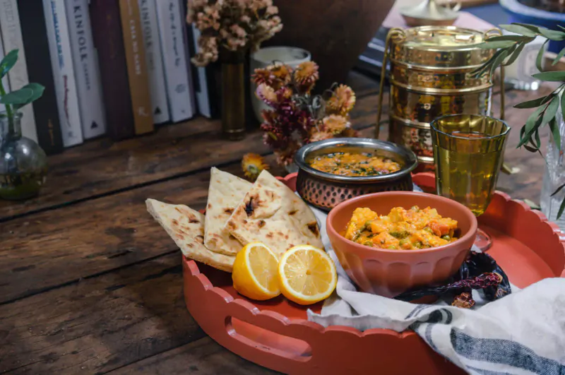 Indian meal on wooden table: naan bread, lentil dal, chickpea curry, lemon wedges on red tray, books and brass tiffin backdrop