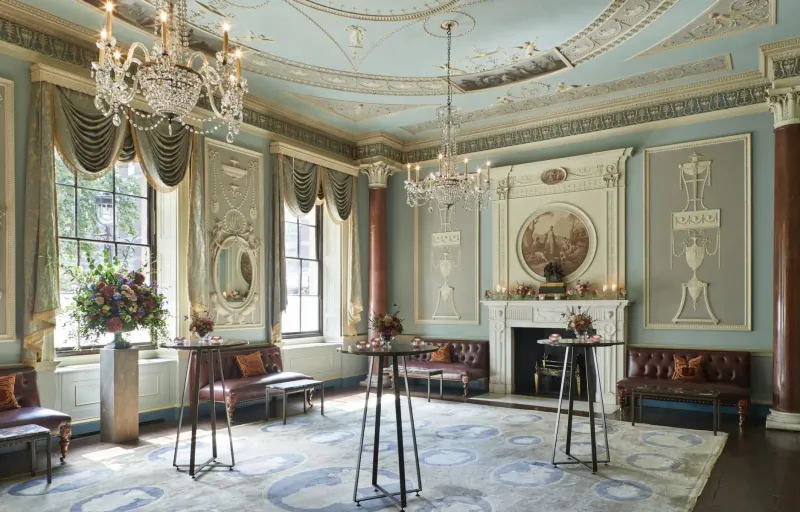 Elegant Home House room with chandeliers, ornate ceiling, high cocktail tables, flowers, and fireplace.