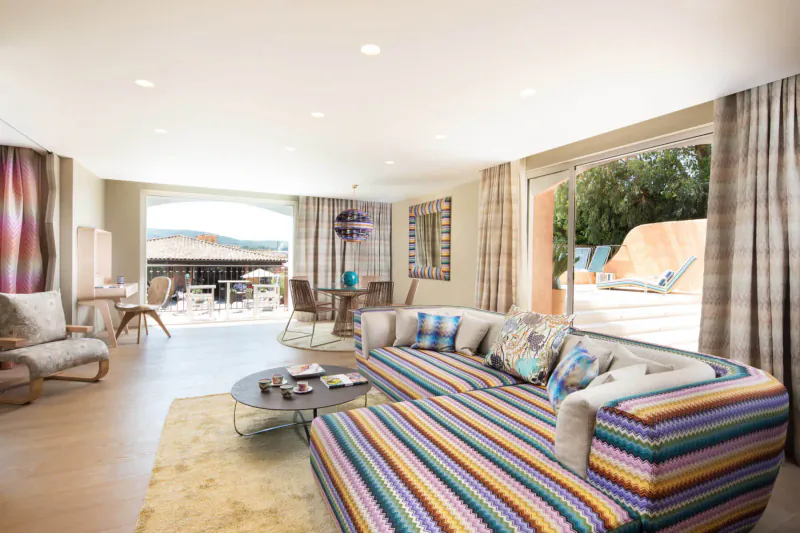 Spacious luxury living room at Hôtel Byblos St Tropez with colorful striped sofa, open doors to pool terrace and garden view.