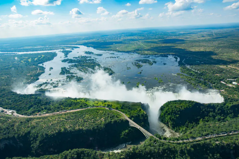 Aerial view of majestic Victoria Falls cascading amid lush green forests and river, under blue sky with clouds.