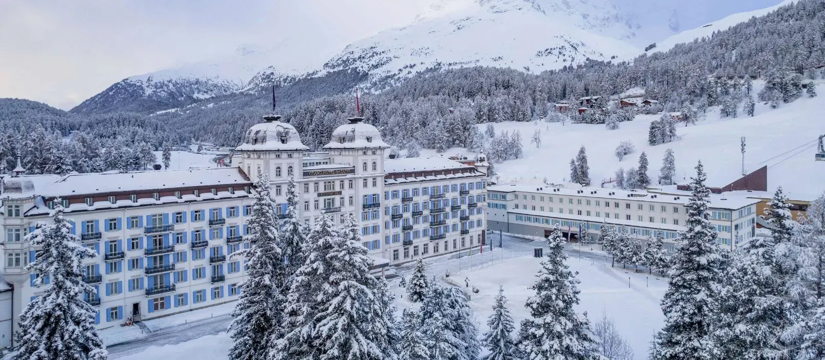Grand Hotel des Bains Kempinski St. Moritz, grand white-and-blue alpine hotel with towers amid snowy mountains and pine trees.
