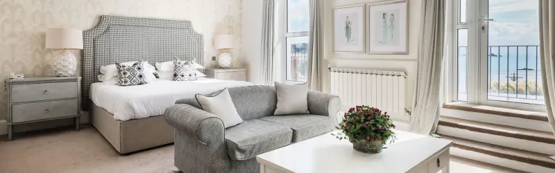 Elegant hotel bedroom with gray upholstered bed, gray sofa, flowers on white table, and sea view balcony.