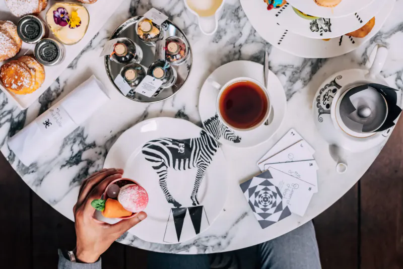 Top-down view of elegant afternoon tea on marble table: zebra plate, pastries, tea, cards, hand holding orange iced drink