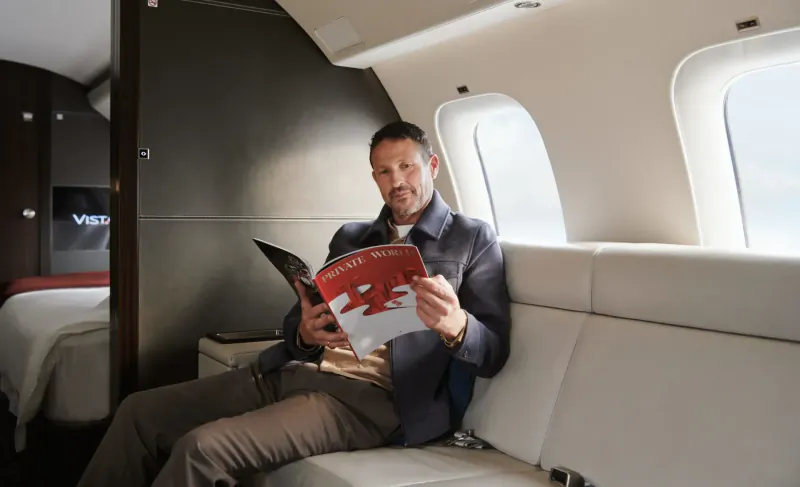 Man in suit reading red VistaJet magazine, relaxed on white leather seat in luxury jet cabin