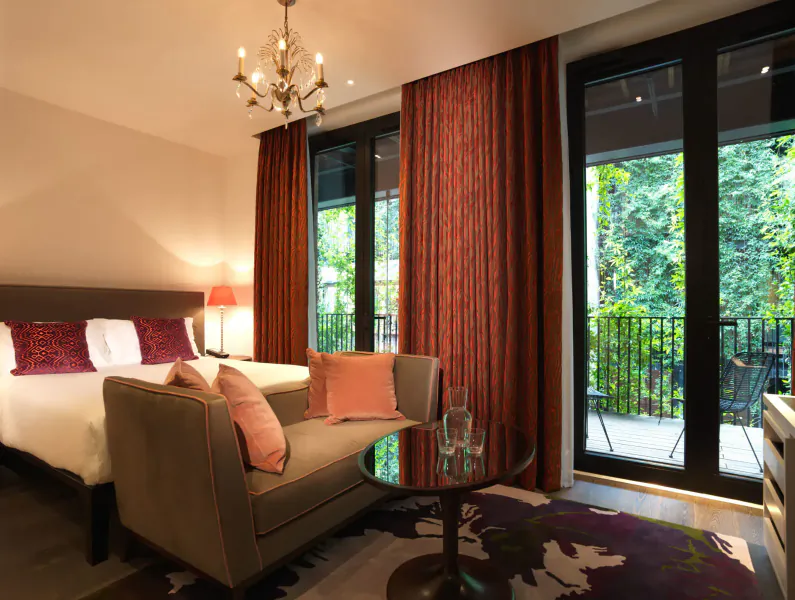 Elegant hotel room at The Mandrake with king bed, pink pillows, loveseat, glass table, chandelier, and garden balcony view.