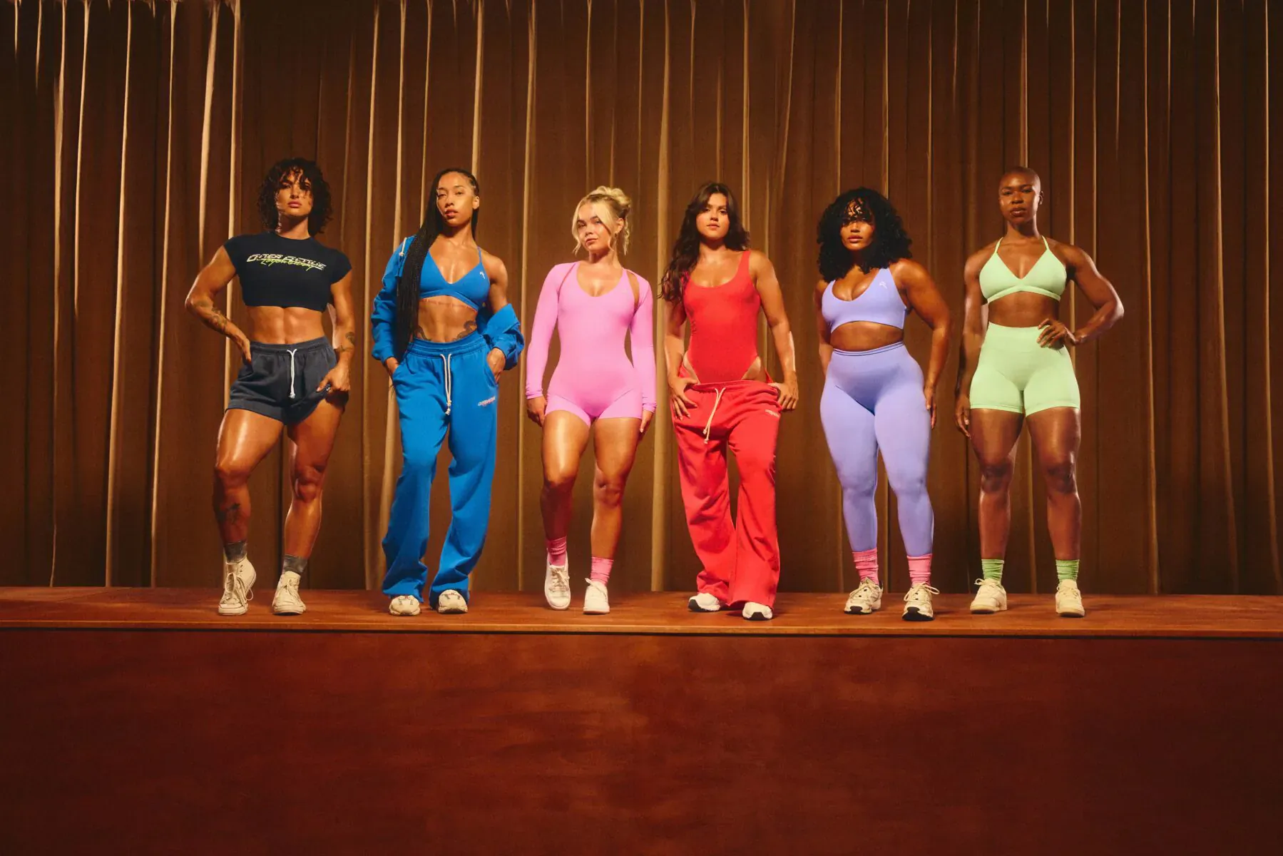 Six diverse women in colorful Oner Active Legendary Capsule outfits posing confidently on stage before gold curtains.